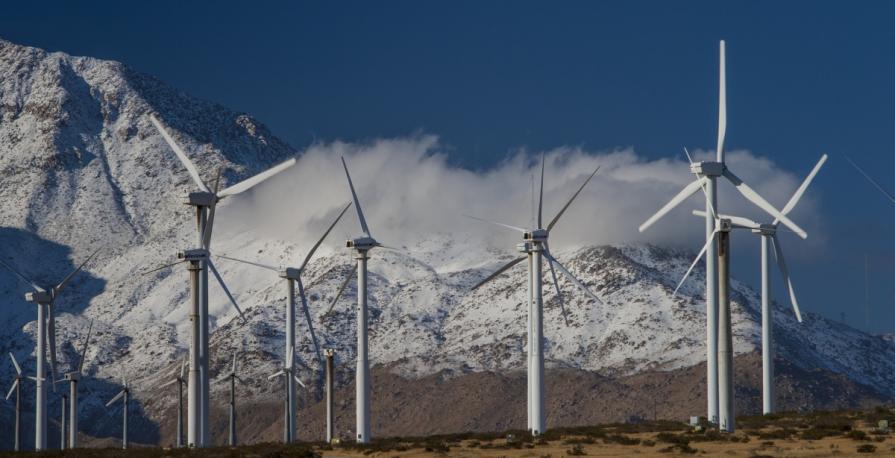 Image shows wind turbines creating energy by actively spinning in front of a background of rocky mountain under a cloudy sky.