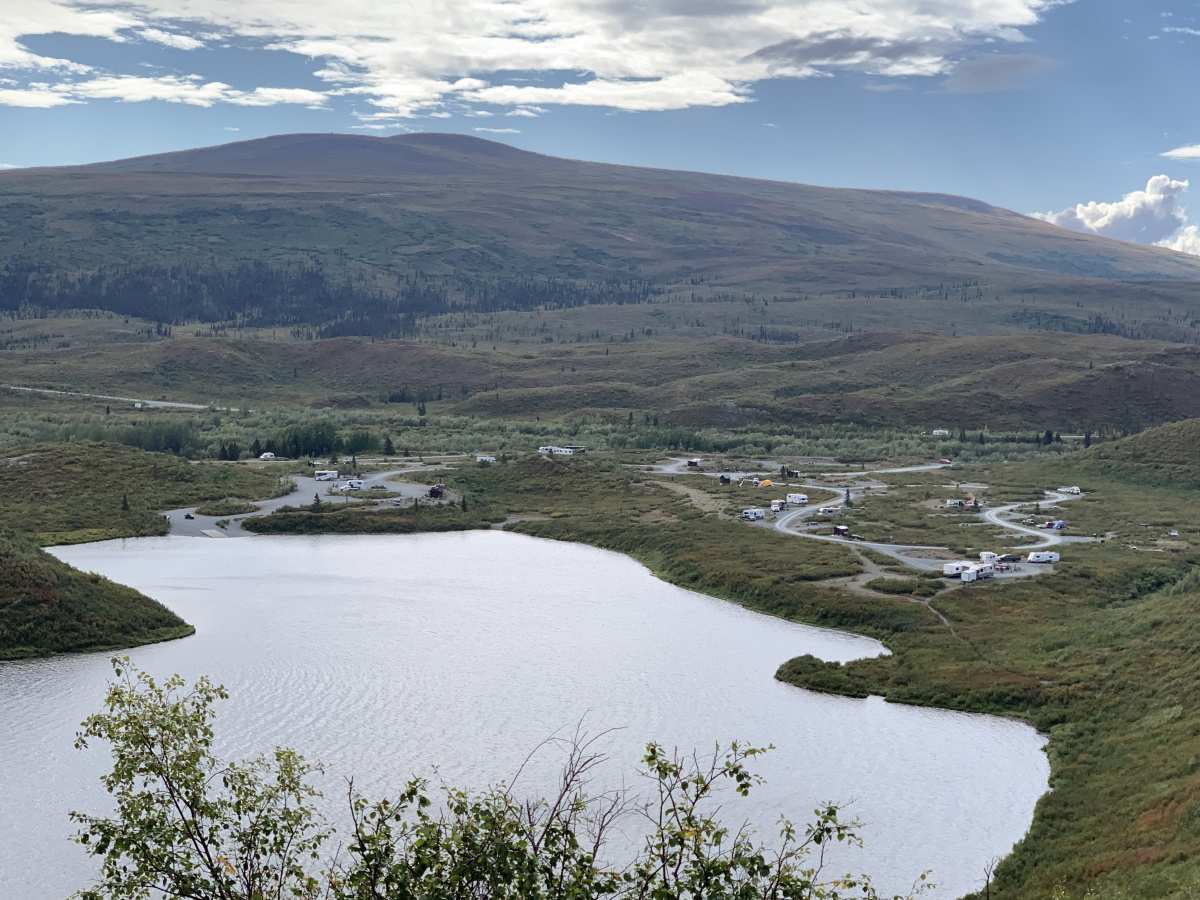 A view of a lake and a campground under blue skies from atop a nearby ridge. There is a mountain in the background.