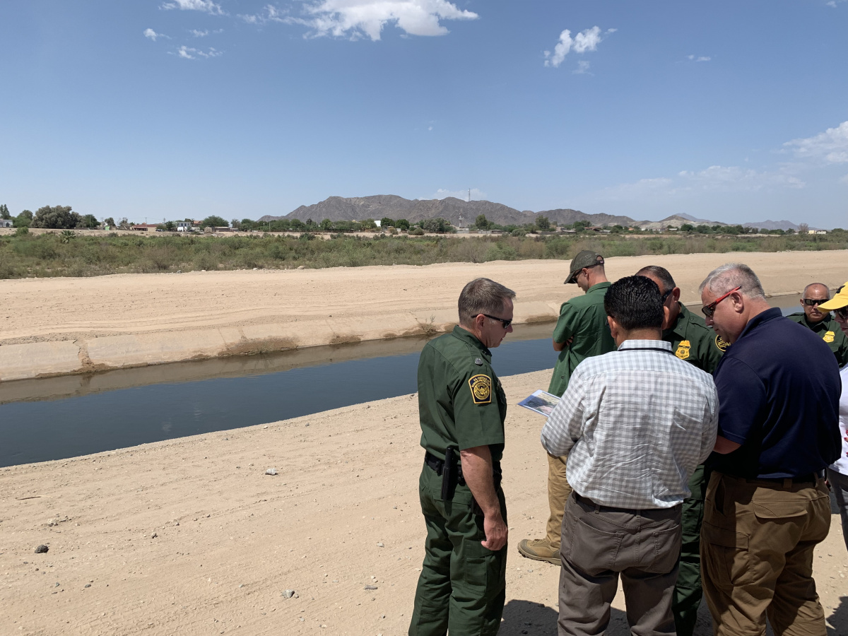 A group of men (some in green uniforms) stand together on a dirt road next to a river.