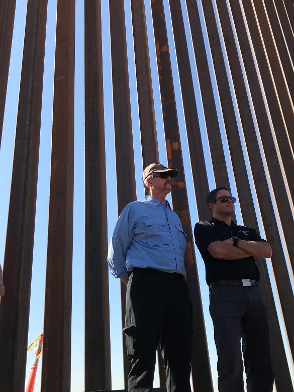 William Perry Pendley standing in front of the border wall with another man.