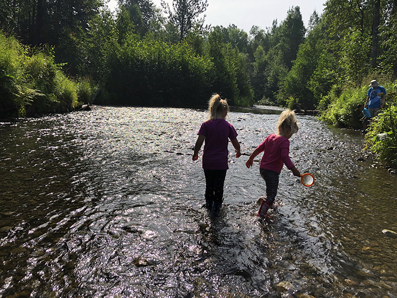 2 girls exploring a shallow creek in water boots with parents on the bank of creek