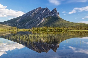 A steep mountain with trees at the base and a blue sky with clouds clearly reflected in a flat lake.