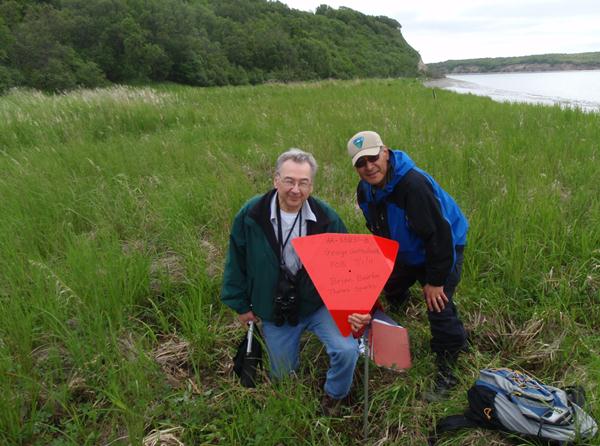 Two men in a grassy field that is bordering a body of water and a hill.