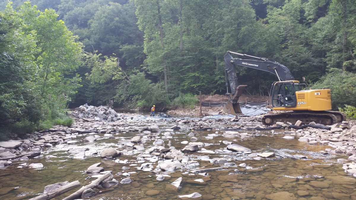 A steam shovel sits on the edge of a rocky stream bank while a worker wearing a hard hat crosses the river in a forest.