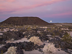 The sky changes color as the moon sets behind a crater formed of ash and cinders; lava rocks and shrubs sit at the base of the crater.  Photo by Bob Wick, BLM.