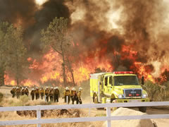 A line of firefighters carry equipment to a fire engine as a wildlife rages in the background. Photo by BLM.