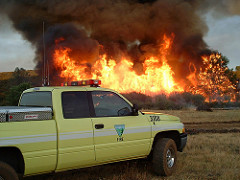 A BLM fire truck sits near a wildfire with smoke and flames shooting high in the sky.  Photo by BLM.