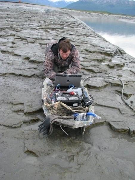 A man kneeling in mud over a sled with computer gear.