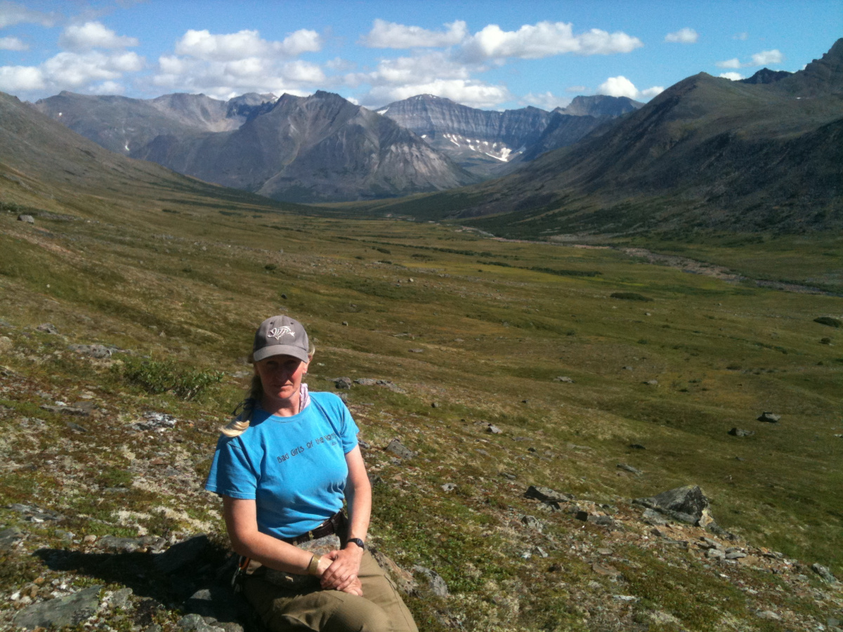 A woman sitting on a mountainside with a valley and more mountains in the background under blue skies