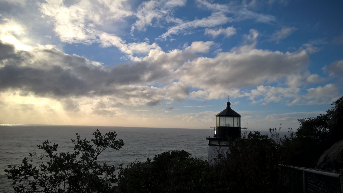 The lighthouse at Trinidad Head.  The lighthouse  overlooks the ocean. The sun shines through the clouds. 