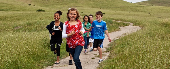 Children running down a path. Fields of green grass gently being blown in the wind surrounds the path. 