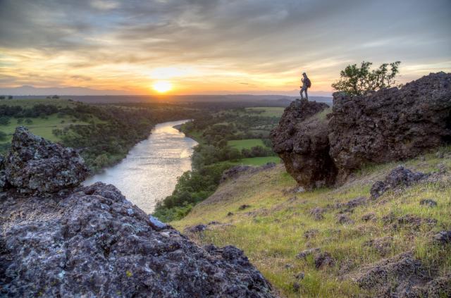 Visitor stands on a rock overlooking sunset over river and green landscape