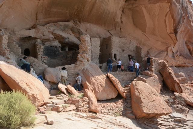 Visitors and Ancestral Lands Conservation Corps workers observe the River House cliff dwelling with a staircase leading down to a rocky area.
