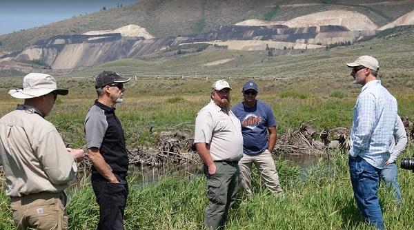 Group of Itafos employees with Habitat Improvement Team members standing in tall green grass on the streambank with the mine operating in the background