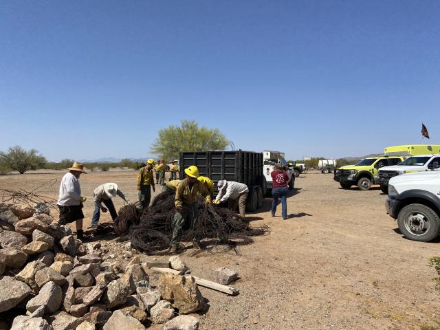 People load barbed wire and other trash into a trailer. There is a big pile of rocks and a large pile of barbed wire just behind the trailer. Trucks are parked off to the side and the sky is blue.