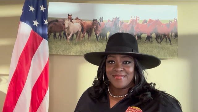 Woman with hat in front of horse picture and near an American flag. 