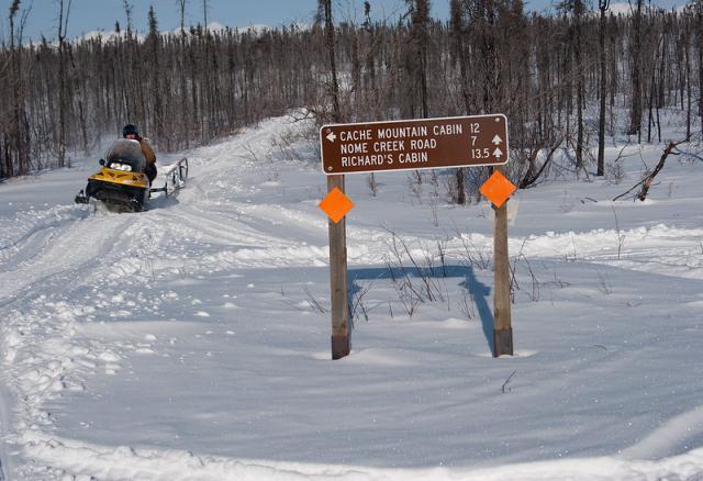 A snowy winter scene: a person on a snow machine pulls a groomer to pack, flatten, and level a winter trail. They are approaching an intersection which is marked by a sign with locations and distances to locations of interest. It is sunny and the rider is surrounded by trees with mountains visible in the distance.