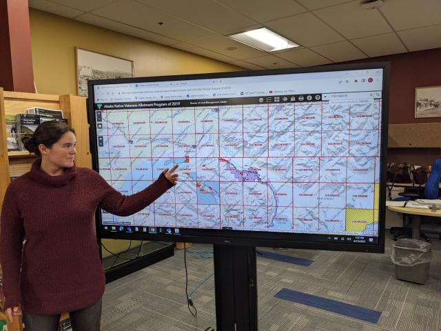 Woman points to a large screen with a map on it. 