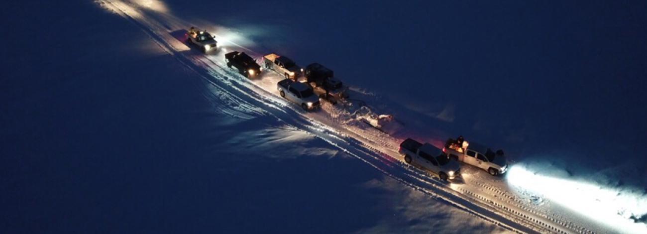Cars and pickup trucks drive the Community Winter Access Trail snow road at night.