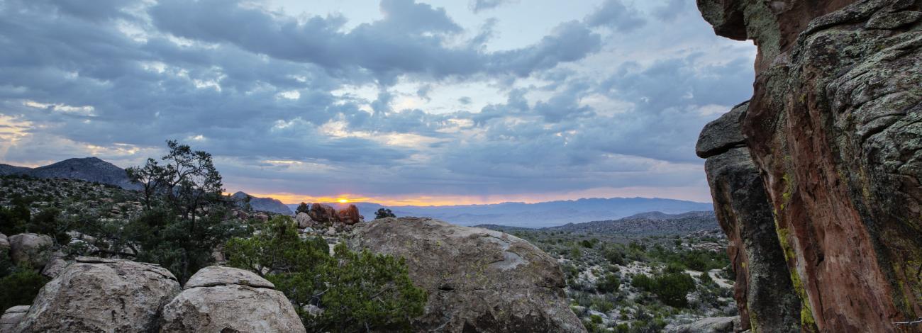 A distant sunset under a sky of soft clouds, frame on the right by a large monolith of stones and smaller rock formations on the left, looking across a verdant Nevada desert landscape
