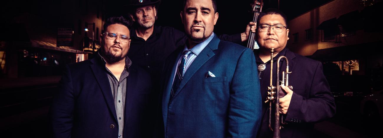 Image of Delberr Anderson and three additional members of the group D'DAT standing outdoors, with some holding instruments including a trumpet, a stand-up bass, and drumsticks.