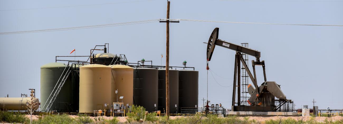Oil pumpjack operating next to large storage tanks in a dry, desert landscape under a clear sky.