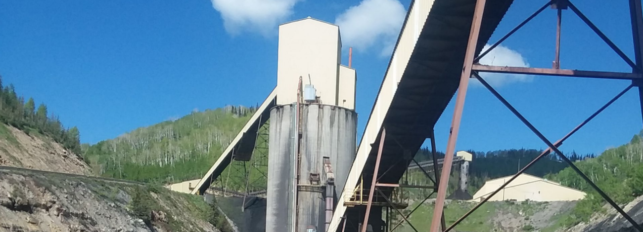 Industrial mining facility with tall silos and conveyor belts set against green hills under a clear blue sky.
