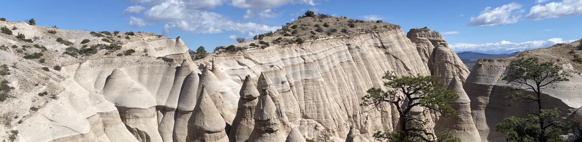 Eroded rock formations and cone-shaped hoodoos rise from a desert landscape under a partly cloudy blue sky, with sparse trees in the foreground.