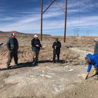 Five individuals standing and one bending over brushing the tracksite. A telephone pole is in the background. The ground is dry, flat, and has little vegetation.