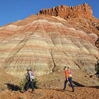 Two people in hiking attire follow a trail along a red, rocky landscape.
