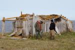 Two men stand in front of a sod home built of drift wood and whale bones with the Arctic ocean in the background