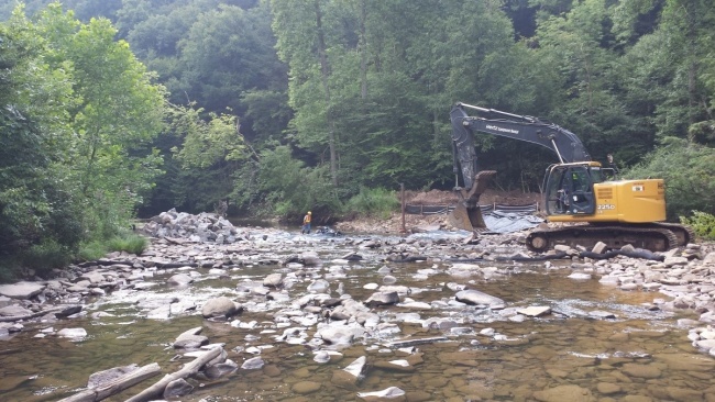 Photo shows heavy equipment in a rock filled stream surrounded by forest trees.