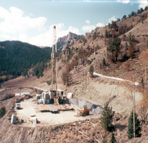 Image shows a flat well pad with a drill rig in an area of dirt on a mountain under a cloudy sky. 