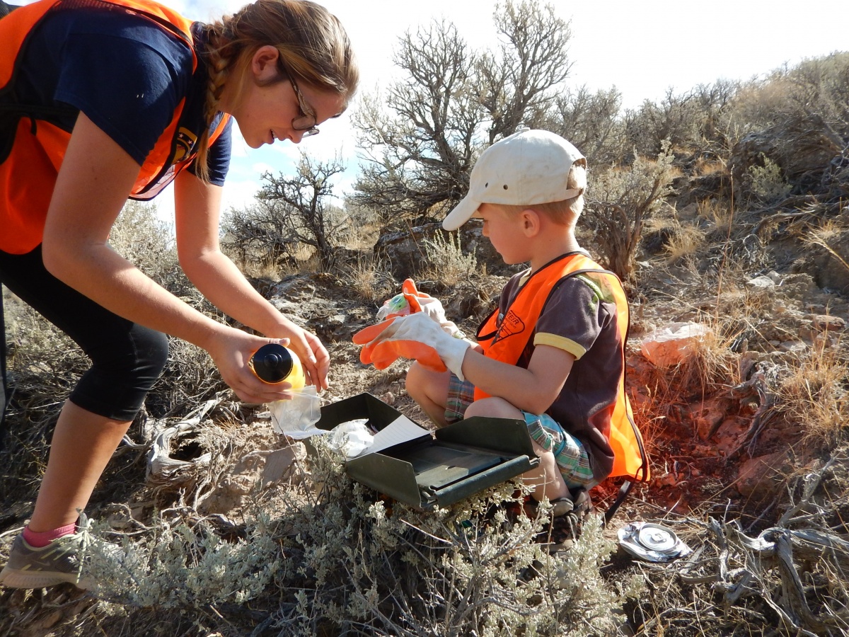 A teenager helps a preschool child open a box on a mountainside.