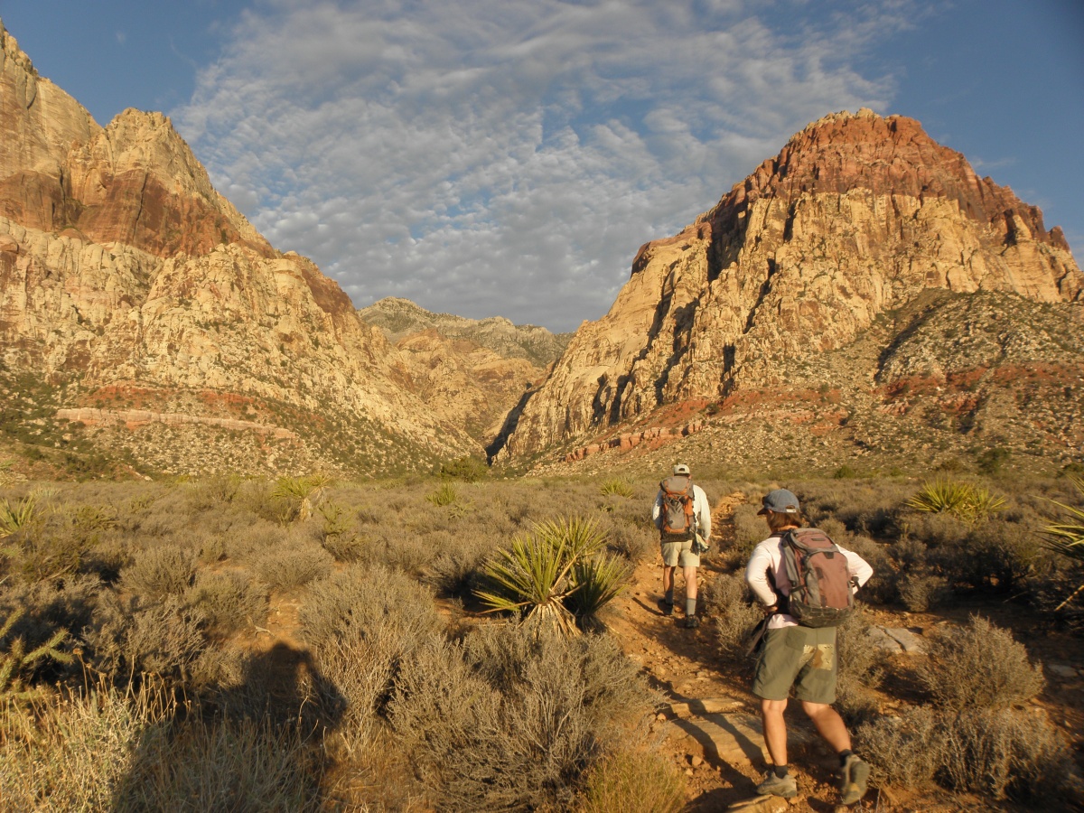 Two hikers in shorts walk through a dramatic red and brown cliffscape at Red Rock Canyons National Conservation Area.