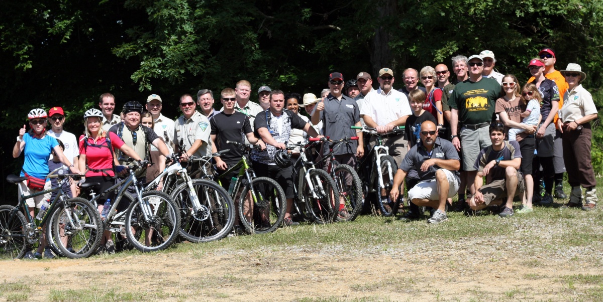 Approximately 35 people pose with their bicycles in front of a stand of trees, near a mountain biking trailhead. 