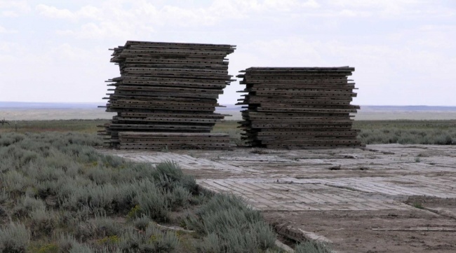 Two piles of brown wooden platforms in a grass field with grey sky in background.