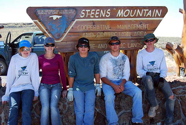 Image of five youth in work clothing sitting on the base of the BLM's Steens Mountain entrance sign.