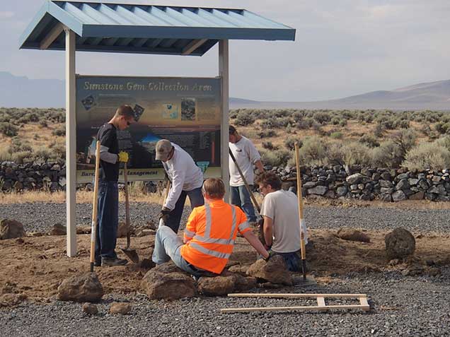 Image of five youth team members in work clothes and holding shovels, digging and spreading dirt at the base of a newly-installed information sign in a gravel area.