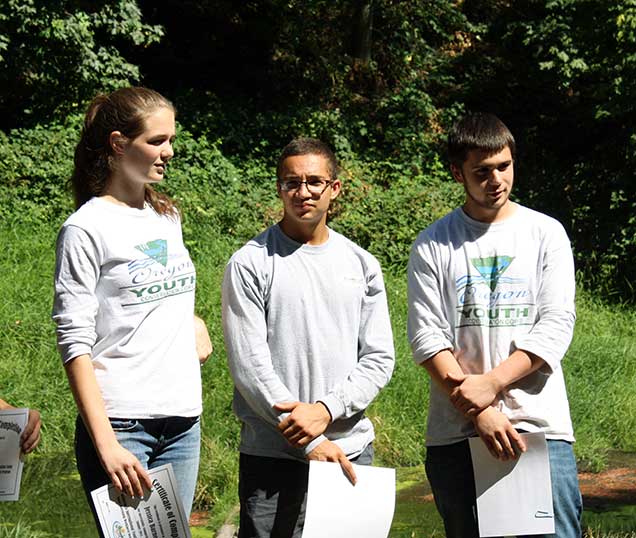 Photo of three young adults with OYCC t-shirts, standing for recognition from a group just off-frame.