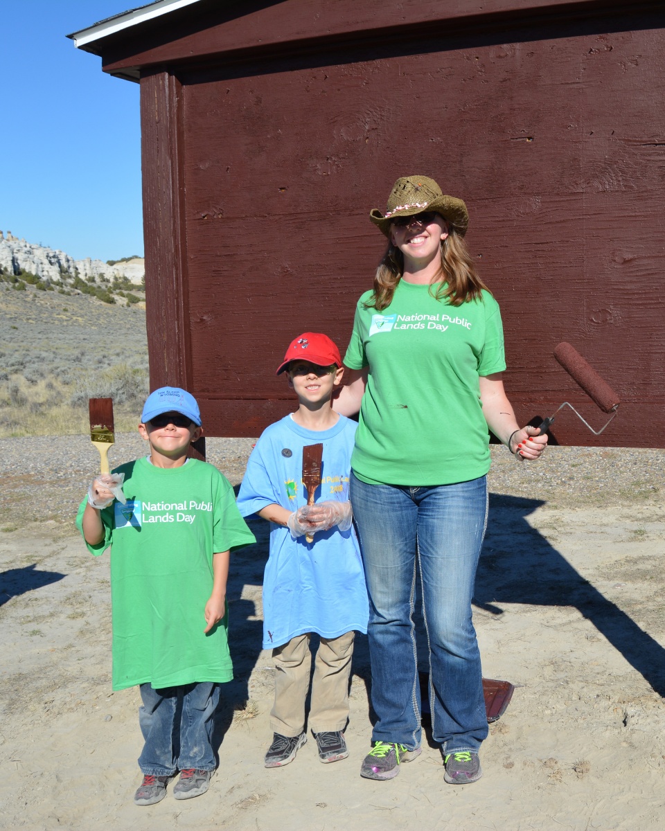 A woman with 2 kids hold paintbrushes and stand next to a building.