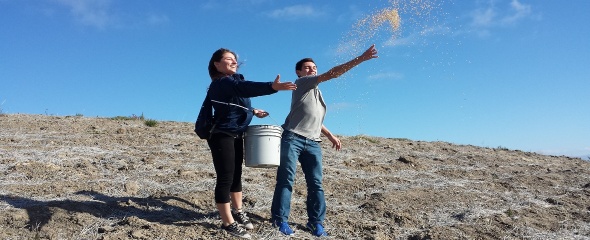Two volunteers spread seeds from a white bucket. They are standing on a brown hill. The sun is shining on their faces as they face into the sun.  The sky is blue with white wispy clouds in the background.