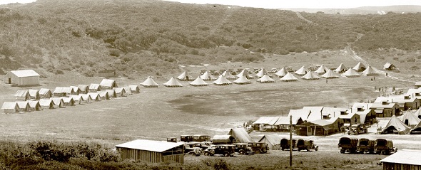 Historic picture of Fort Ord Army Base.  The picture is sepia colored.  In the foreground there is a small wooden building. Old vehicles are parked along a small road. In the background are rows of tents pitched in rows before a small hill. 