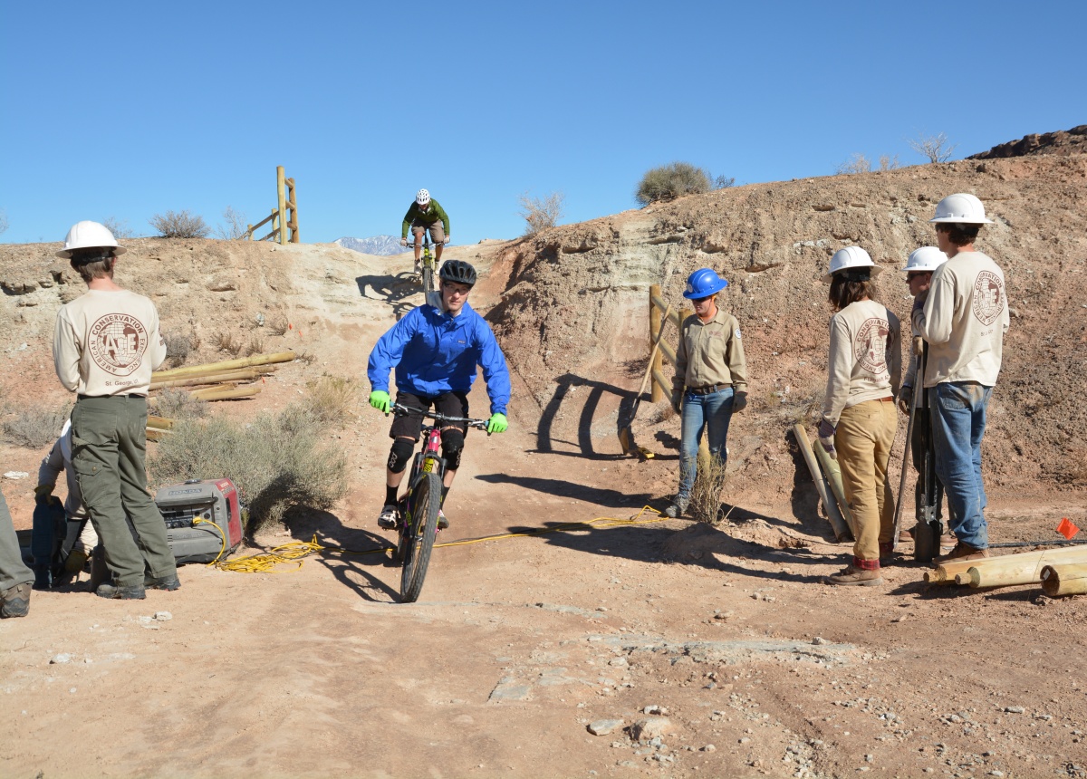 Two mountain bikers ride the Bear Claw Poppy Trail while ACE trail crew members work to build a fence.