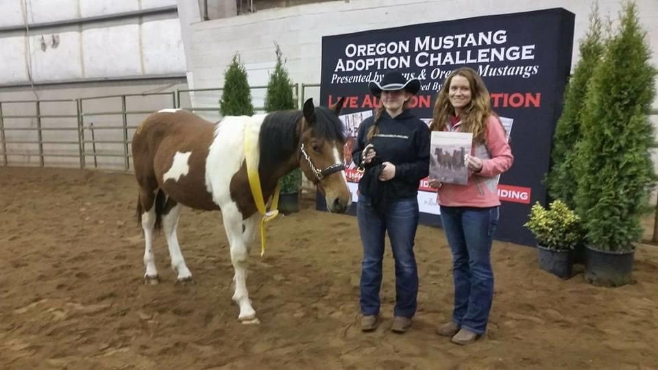Two people standing with a horse in from of a banner that reads Oregon Mustang Challenge