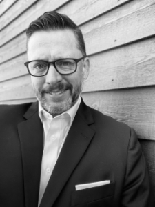 A black and white photo of a man with short dark hair and glasses wearing a suit jacket leaning against a wall