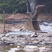 Photo shows heavy equipment in a rock filled stream surrounded by forest trees.