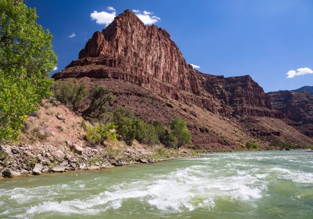 A green-tinted river with rapids flows past a large red rock or hill with the rocky bank and greenery on the side.