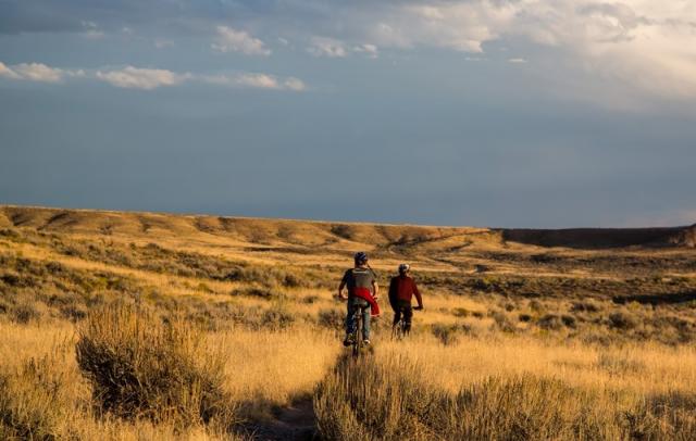 Two mountain bikers riding on trails within the McCoy Flats Trail System managed by the Vernal Field Office. Grass is all around them and clouds dot the sky.