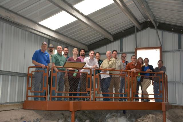 Many people stand close together on a metal walkway overlooking a fossil bed inside a building at Jurassic National Monument.